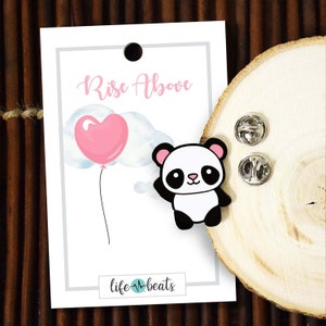 May include: A black and white panda bear cartoon enamel pin with pink ears. The pin is on a wooden surface with a white card behind it. The card has the text "Rise Above" and "life-beats" on it. The card also has a pink heart shaped balloon with a white cloud behind it.