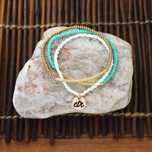 May include: Three beaded bracelets in shades of gold, turquoise, and white are displayed on a light gray stone. One bracelet has a gold bar design. A gold charm with the letters "ctr" hangs from one bracelet.