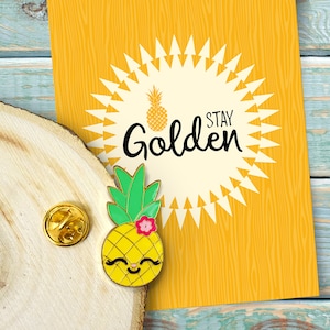May include: A yellow pineapple enamel pin with a smiling face and pink flower. The pin is on a wooden surface with a yellow card in the background. The card has a white circle with a pineapple and the text "Stay Golden".