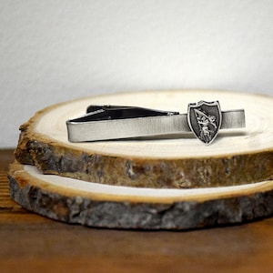 May include: A silver-toned tie clip with a brushed metal finish. The clip features a decorative shield emblem with a detailed figure. The tie clip is displayed on a rustic wooden surface, showcasing its design and craftsmanship.