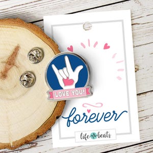 May include: A blue enamel pin with a white hand signing "I love you" in American Sign Language. The pin has a pink heart and a pink banner that says "LOVE YOU!" The pin is on a wooden surface with a white card that says "forever" in cursive and "life beats" in a pink heart graphic.