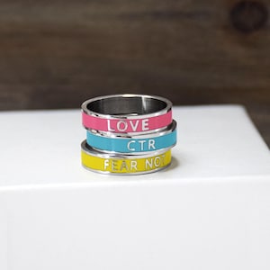 May include: Three stacked rings with colorful enamel bands. The top ring is pink with the word "LOVE", the middle ring is blue with "CTR", and the bottom ring is yellow with "FEAR NOT". The rings are made of silver-colored metal.