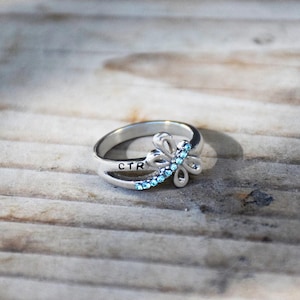May include: A silver ring with a delicate flower design featuring blue crystals. The ring has the inscription "CTR" on the band.