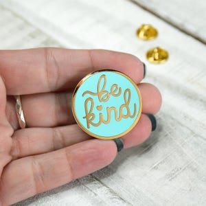 May include: A gold-colored enamel pin with a light blue background. The pin features the words "be kind" in a gold-colored script font with a small heart symbol.