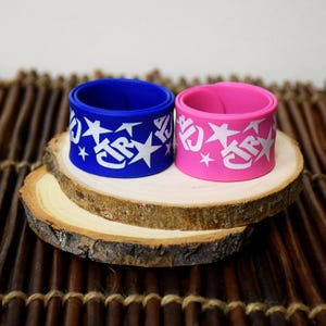 May include: Two colorful wristbands, one blue and one pink, are displayed on a wooden slice. Each band features the text "JTR" surrounded by white stars. The bands are cylindrical and appear to be made of a flexible material.