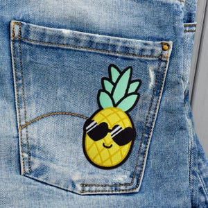May include: A yellow pineapple with a green leafy top and black sunglasses is sewn onto a blue denim pocket.