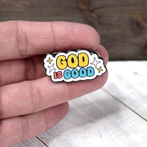 May include: A cloud-shaped enamel pin with a black outline featuring the yellow text GOD and the blue text IS GOOD. The design includes small four-pointed star accents in yellow and white, set against a white background within the cloud shape.