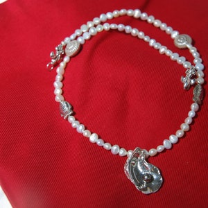 Puede incluir: Un collar de perlas blancas con colgantes de plata en forma de concha y estrella de mar. El collar está sobre un fondo de tela roja.