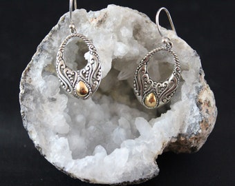 May include: A pair of silver earrings with a gold accent in the center of each earring. The earrings are oval shaped and have a delicate, intricate design. The earrings are displayed on a white rock.