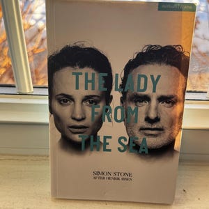 Può includere: Un libro intitolato "THE LADY FROM THE SEA" di Simon Stone, da Henrik Ibsen. La copertina presenta due volti, uno femminile e uno maschile, con il titolo in lettere turchesi. Il libro è appoggiato su un davanzale.