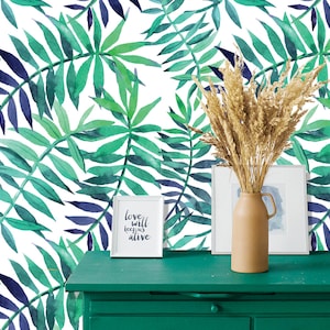 Puede incluir: Una cómoda verde con un fondo blanco con un patrón de acuarela de hojas verdes y azules. La cómoda tiene una impresión enmarcada con el texto "love will keep us alive" y un jarrón con trigo seco.