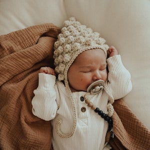 May include: A baby wearing a white onesie and a beige crocheted hat with a pacifier in their mouth. The baby is lying on a white surface with a brown blanket.