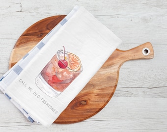 May include: White kitchen towel with blue stripes folded on a wooden cutting board. The towel features a watercolor illustration of an old fashioned cocktail with a cherry and orange slice. The text "CALL ME OLD FASHIONED" is printed on the towel.