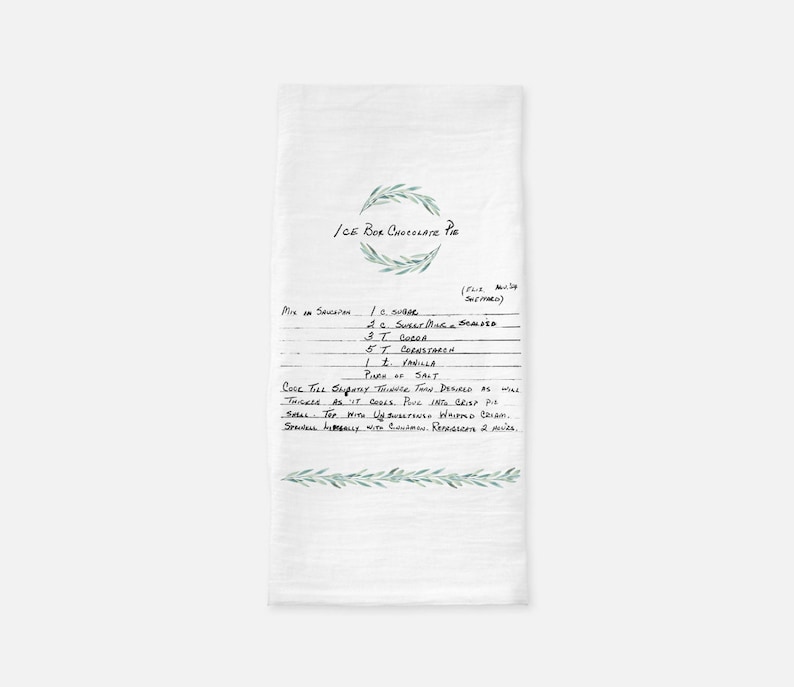 May include: A white kitchen towel with a handwritten recipe for Ice Box Chocolate Pie. The recipe includes ingredients like sugar, cocoa, cornstarch, vanilla, and salt. The towel is decorated with a green olive branch design.