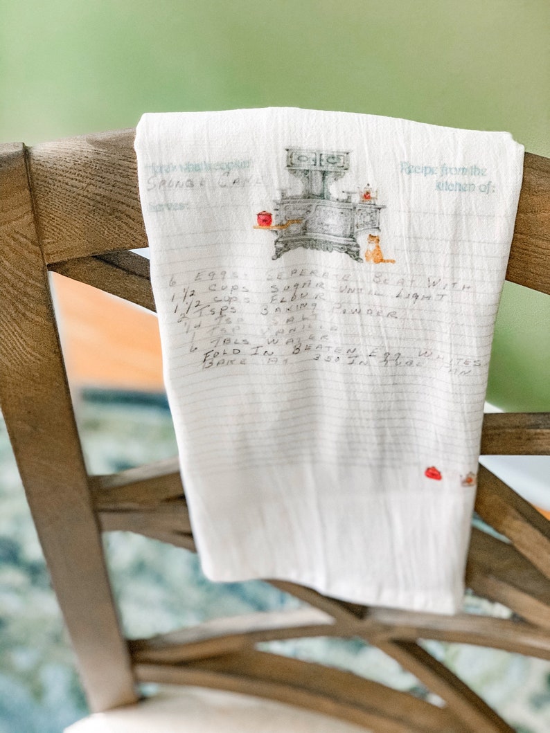 May include: A white tea towel with a vintage stove and cat illustration. The towel has handwritten text that reads "Recipe from the kitchen of:" and a recipe for "Sponge Cake" with ingredients and instructions.
