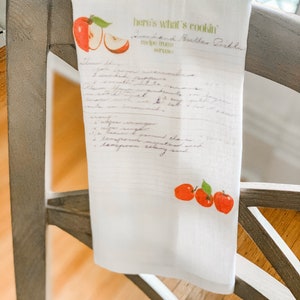 May include: A white kitchen towel with a handwritten recipe for "Greenhand Butter Pickles" and red apple illustrations. The recipe includes ingredients like "greenhand cucumbers", "red pepper", "sugar", "salt", "mustard seed", "onion", "garlic", "vinegar", "sugar", "ground cloves", "celery seed", "mustard seed", and "ground ginger".