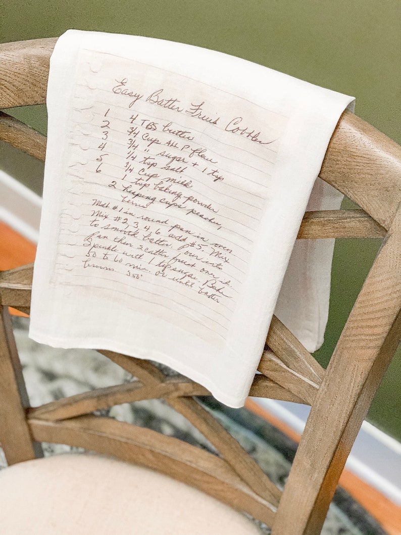 May include: A handwritten recipe for Easy Batter Fruit Cotter on a white kitchen towel hanging on a wooden chair. The recipe includes ingredients such as butter, all-purpose flour, sugar, salt, milk, baking powder, and peaches.