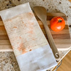 May include: A white tea towel with a handwritten spaghetti recipe in brown ink. The recipe is stained with orange and yellow splotches. The tea towel is folded and lying on a wooden cutting board with a wooden spoon and a red tomato.