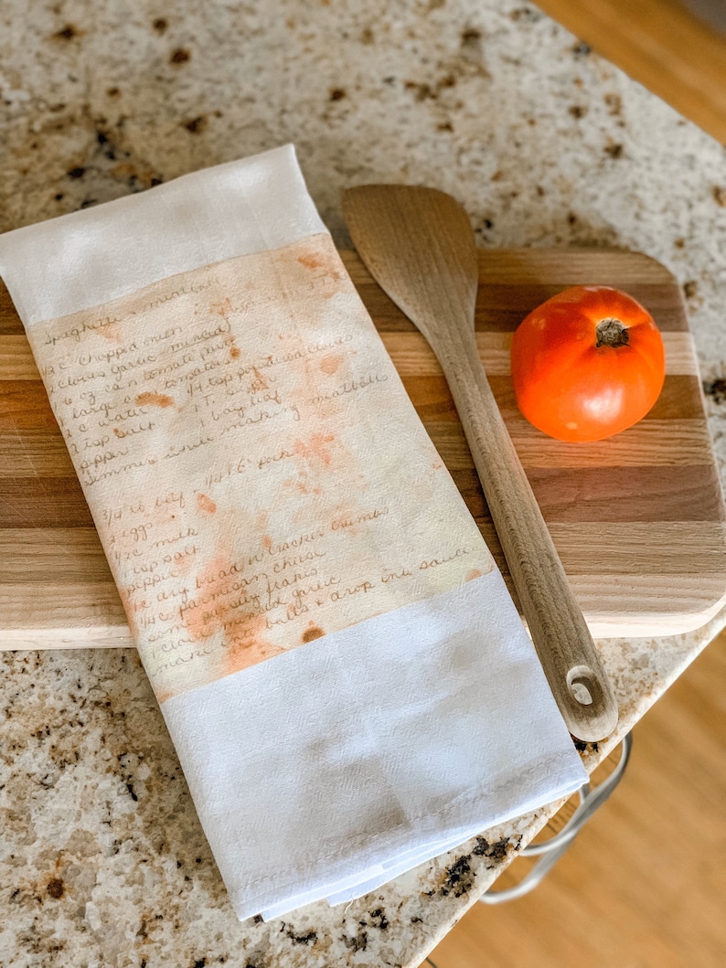 May include: A white tea towel with a handwritten spaghetti recipe in brown ink. The recipe is stained with orange and yellow splotches. The tea towel is folded and lying on a wooden cutting board with a wooden spoon and a red tomato.