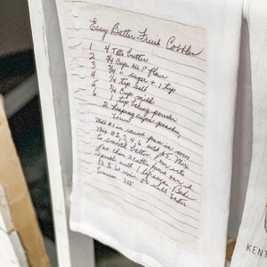 May include: A handwritten recipe for Easy Batter Fruit Cobbler on a white tea towel. The recipe lists ingredients including butter, all-purpose flour, sugar, salt, milk, baking powder, and peaches.
