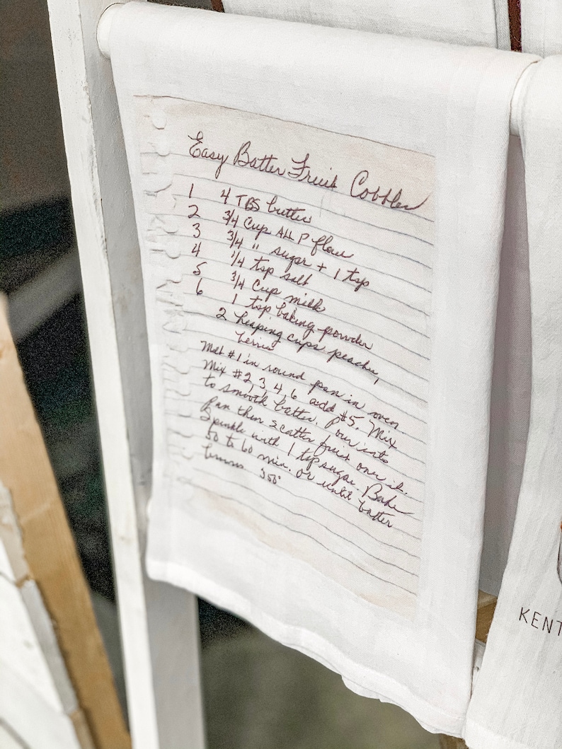 May include: A handwritten recipe for Easy Batter Fruit Cobbler on a white tea towel. The recipe lists ingredients including butter, all-purpose flour, sugar, salt, milk, baking powder, and peaches.