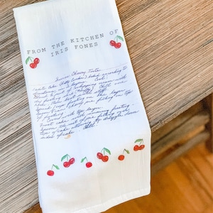 May include: A white kitchen towel with a handwritten recipe for a cherry torte. The recipe is from "The Kitchen of Iris Fones". The towel is decorated with red cherries.