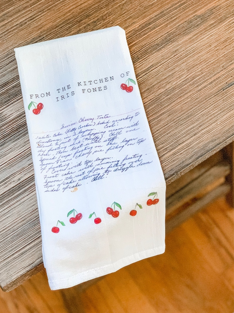 May include: A white kitchen towel with a handwritten recipe for a cherry torte. The recipe is from "The Kitchen of Iris Fones". The towel is decorated with red cherries.
