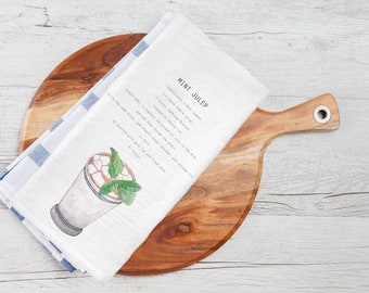 May include: White kitchen towel with blue stripes, folded on a wooden cutting board. The towel features a watercolor illustration of a mint julep cocktail and a recipe for the drink: "MINT JULEP INGREDIENTS: 1. 2 oz. Bourbon 2. 1/2 ounces simple syrup 3. 8-10 mint leaves Crushed or muddled DIRECTIONS: 1. Muddle the mint with the simple syrup and bourbon in the bottom of the glass. 2. Fill the glass with crushed ice and top with more syrup. 3. Garnish with a sprig of mint.  Enjoy!"