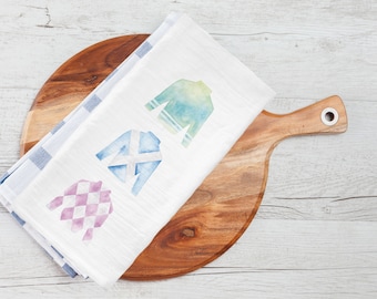 May include: A white kitchen towel with three watercolor images of horse racing silks. The silks are blue and white, green and white, and pink and white.