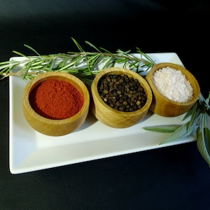 May include: A white rectangular plate holds three small wooden bowls filled with spices. One bowl contains red paprika, another black peppercorns, and the third coarse salt. Fresh rosemary and sage sprigs are arranged around the bowls, creating a culinary still life.