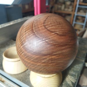 Op de afbeelding: Een glad, bolvormig houten object met een rijke, donkerbruine houtnerf, rustend op een lichtgekleurde, ronde houten voet. Een apart, kleiner, komvormig houten stuk staat aan de linkerkant van de bol.