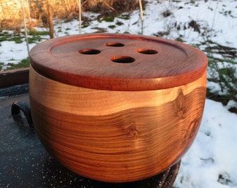 May include: A round wooden container with a smooth, polished finish featuring a prominent wood grain pattern. The lid is shaped like a large button with four circular holes in the center, showcasing a deep reddish-brown hue and natural wood tones.