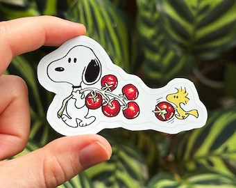 May include: A white sticker featuring Snoopy holding a bunch of red cherries, with Woodstock chasing a single cherry. The sticker has a white border and is held against a background of green leaves.