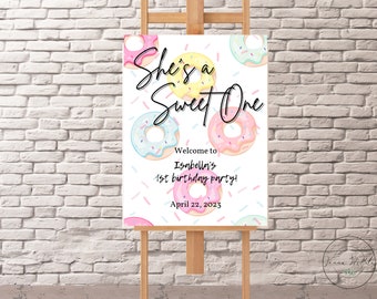 Könnte beinhalten: Ein weißes Schild mit Donut-Motiv für eine erste Geburtstagsfeier. Das Schild sagt "She's a Sweet One" mit bunten Donuts und Streuseln. Der Text sagt auch "Willkommen zu Isabellas 1. Geburtstagsparty! 22. April 2023".