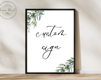 Op de afbeelding: Een wit, ingelijst bord met de woorden "custom sign" in elegant zwart schrift. Het bord is versierd met aquarel groen in de hoeken. De lijst is zwart. Het bord wordt weergegeven op een houten oppervlak.