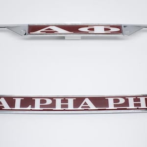 以下が含まれることがあります： クロム製のナンバープレートフレーム。マホガニー色の背景に白い文字で「ALPHA PHI」と書いてあります。
