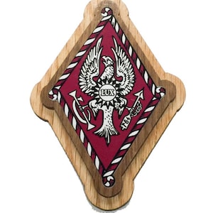 May include: A wooden plaque with a red and white diamond-shaped design. The diamond features a white eagle with a sunburst and the word "LUX" in a circle. The eagle is holding a key and an anchor. The plaque is surrounded by a wooden frame.