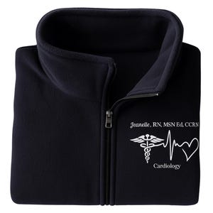 May include: A navy blue fleece zip-up jacket with a high collar. The jacket features a white caduceus, heartbeat, and heart graphic with the word "Cardiology" and the name "Jeanelle, RN, MSN Ed, CCRN".