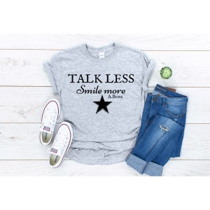 Pode incluir: Uma camiseta cinza com o texto "TALK LESS Smile more A.BURR" e uma estrela preta impressa na frente. A camiseta está colocada sobre uma superfície de madeira com um par de tênis brancos e uma calça jeans azul.