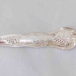 May include: A silver serving fork with three tines. The handle features an ornate design with a cluster of grapes. The fork is made of a shiny, reflective metal, likely silver or a silver-colored alloy. The fork is designed for serving.