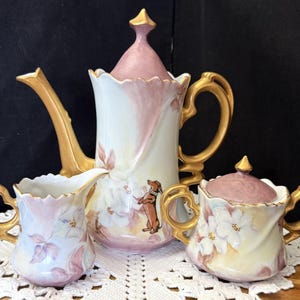 May include: A vintage porcelain tea set featuring a teapot, creamer, and sugar bowl. The set is painted with a floral design in shades of pink, white, and yellow, with gold accents. A dachshund dog is painted on the teapot. The set is displayed on a white lace doily.