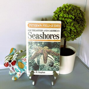 May include: A paperback book titled Peterson Field Guides Southeastern and Caribbean Seashores by Eugene H. Kaplan, featuring a cover image of a starfish on a shell, displayed on a silver metal stand with a white background.