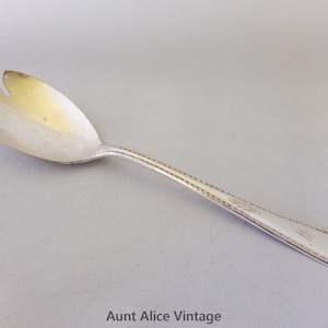 May include: Silver serving spoon with a three-pronged head. The handle has a detailed pattern and a decorative end. The spoon is likely vintage and is designed for serving salads or other dishes. The spoon is made of a shiny metal.
