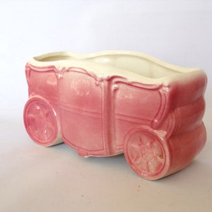 May include: A pink ceramic planter shaped like a horse-drawn carriage. The planter has white accents and is in the shape of a carriage with four wheels.