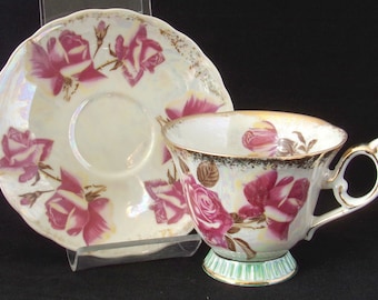 May include: A white teacup and saucer set with a pink and white rose pattern. The cup and saucer have a gold rim and a pearlescent finish.