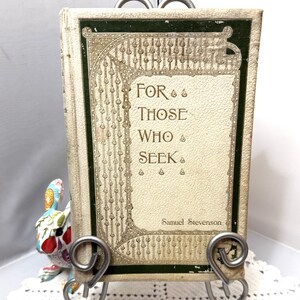 May include: A vintage book titled "For Those Who Seek" by Samuel Stevenson, displayed on a decorative metal stand. The book has a cream-colored cover with green and gold decorative accents. A colorful ceramic bunny figurine is visible to the left.