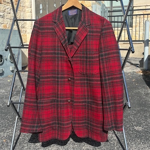 May include: A red and black plaid blazer hanging on a wooden hanger. The blazer has a classic design with a notched lapel and three buttons. The jacket is displayed on a metal drying rack outdoors.