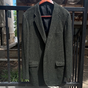 Könnte beinhalten: Ein dunkelgrüner Blazer mit Zwei-Knopf-Verschluss, der an einem Holzbügel hängt. Der Blazer hat zwei Vordertaschen und ein fallendes Revers. Der Stoff scheint eine strukturierte Wolle oder Wollmischung zu sein.