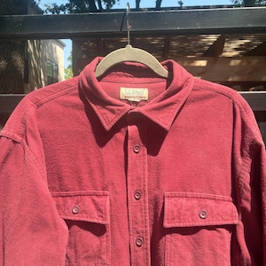 May include: A burgundy button-down shirt with two chest pockets. The shirt has a collared neckline and is hanging on a hanger. The brand label "L.L. Bean" is visible on the collar.