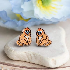 Peut inclure: Une paire de boucles d'oreilles en bois représentant un singe doré à nez retroussé de dessin animé. Les boucles d'oreilles ont une fourrure orange, un ventre blanc et des yeux bleus. Le singe est assis, les jambes croisées.